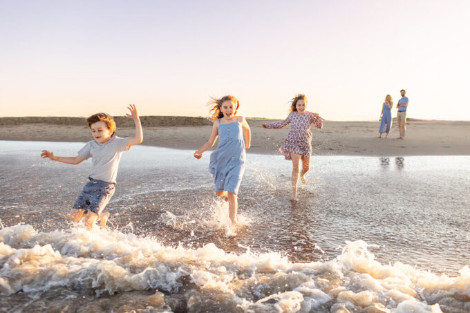 three siblings run wildly and happily into the ocean waves giggling together and their parents stand back and watch enjoying their time spent together as the sun sets on the beach during a family photography session in virginia beach.