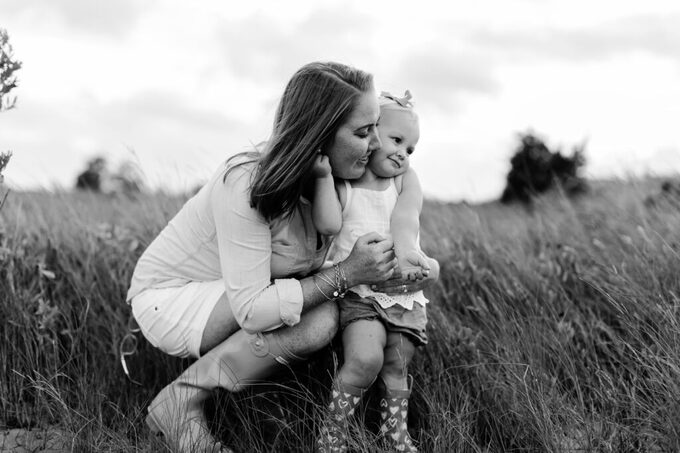 emotional black and white photo of a mom and her baby girl snuggled up together in a warm embrace in a grassy field wearing rain boots together in a warm hug during a playful mother and daughter photo session.