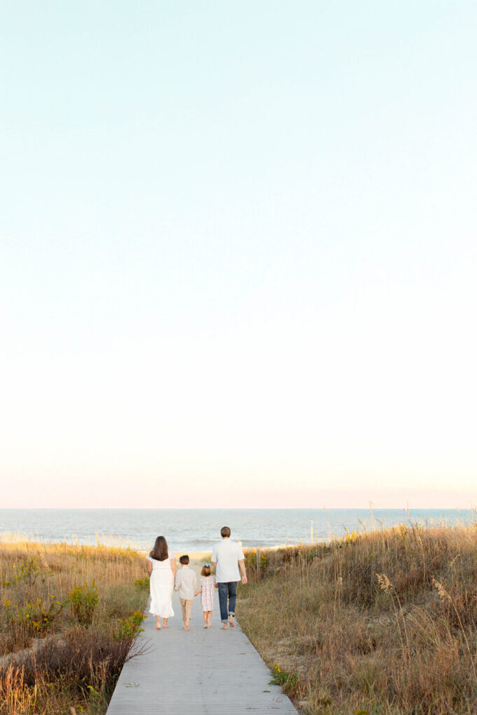 beautiful family of four walk hand in hand down the boardwalk together towards the ocean during sunset dressed in beautiful neutral colors during a relaxed, playful family session in virginia beach.