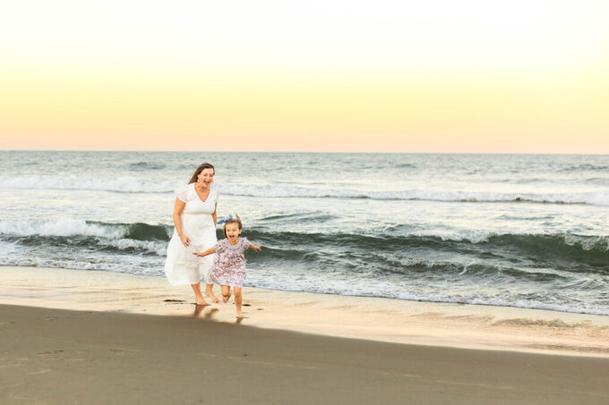 Mother and daughter in beautiful neutral colors laughing and playing candidly in the shore break at sunset during a play filled family session in Virginia Beach