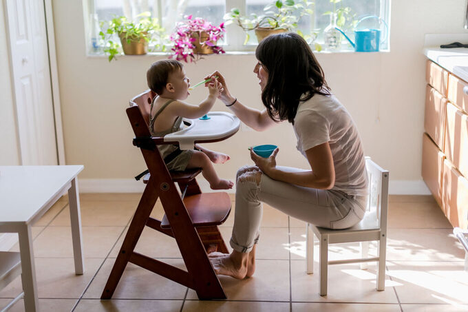 Mother and toddler son sitting in the kitchen in beautiful sun drenched connecting together and the mom feeds her son spoonfuls of baby food in a beautiful at home session filled with love.